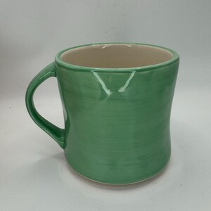 May include: A handmade ceramic mug with a rounded shape and a green exterior. The mug has a curved handle and a cream-colored interior. The green glaze has visible brushstrokes, adding to its unique, artisanal appeal.