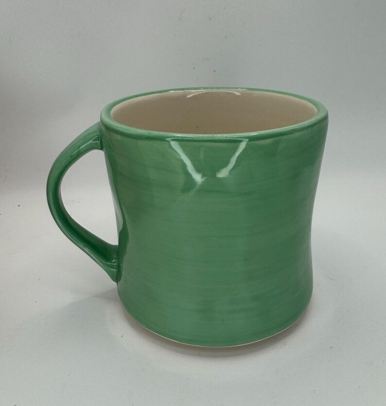 May include: A handmade ceramic mug with a rounded shape and a green exterior. The mug has a curved handle and a cream-colored interior. The green glaze has visible brushstrokes, adding to its unique, artisanal appeal.