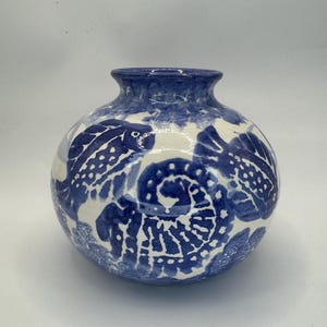 May include: A round ceramic vase with a glossy white finish and hand-painted deep blue designs. The surface features stylized illustrations of fish and a large spiral shell pattern, with a solid blue rim around the circular opening at the top.
