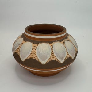 May include: A round, terracotta-colored ceramic pot with a wide opening. The pot features a brown band with white, leaf-shaped designs and a cream-colored border. The base and rim are a lighter shade of terracotta.