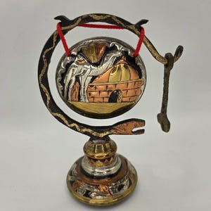 May include: A decorative metal gong on a stand featuring a circular plate with a camel and building relief in silver, copper, and brass tones. The stand is dark metal with a curved arm and a red string holding the gong, accompanied by a small striker.