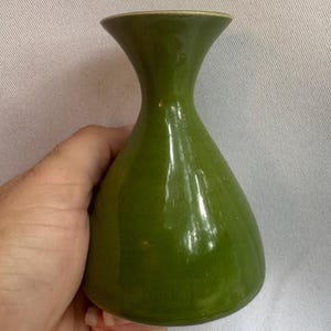 May include: A small, glossy olive green ceramic vase with a smooth, rounded body that tapers into a narrow neck and flares out at the top opening. The surface has a reflective finish and a consistent, solid green color throughout the entire piece.