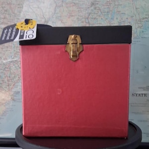 May include: A square storage box with a red base and a black lid. The box features a central gold-toned metal latch on the front. A white tag with black text is attached to the top left corner of the lid, displaying the brand name COVA.