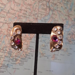 May include: A pair of gold-toned hoop earrings with a glittery finish. Each earring features a large, faceted, deep pink gemstone and is encrusted with clear, sparkling crystals. The earrings are displayed on a black stand.