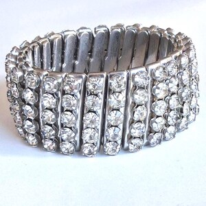 May include: A silver-toned stretch bracelet with multiple rows of clear, sparkling rhinestones. The bracelet is designed with rectangular metal links that allow for flexibility and expansion. The rhinestones are set closely together, creating a dazzling effect.