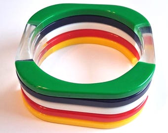May include: A square bangle bracelet with a clear, transparent outer layer. The bracelet features layers of green, navy, white, red, and yellow. The bracelet is made of a glossy, colorful material.