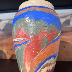 Könnte beinhalten: Eine handgefertigte Keramikvase mit strukturierter Oberfläche. Die Vase hat eine Farbpalette aus Rot, Blau, Orange und Grün mit einem cremefarbenen Rand. Die Farben sind verwirbelt und vermischt, wodurch ein einzigartiges, künstlerisches Design entsteht.