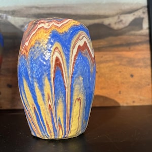 Könnte beinhalten: Kleine, handgefertigte Keramikvase mit einem einzigartigen, farbenfrohen Design. Die Vase weist eine Mischung aus blauen, gelben, roten und weißen vertikalen Streifen auf, die einen Marmoreffekt erzeugen. Die Vase hat einen abgerundeten Körper und eine leicht ausgestellte Oberseite.