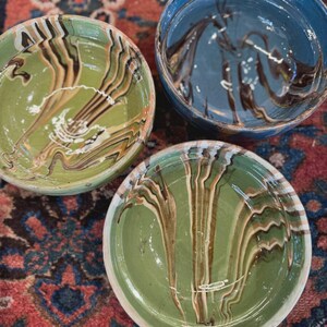 May include: Three ceramic bowls with a marbled design in shades of green and blue. The bowls have a glossy finish and a white rim. The bowls are arranged on a patterned rug with red, blue, and brown colors.