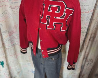 May include: A red varsity jacket with the letters "PK" in white and black on the front. The jacket has a black, white, and red striped collar and cuffs. The jacket is buttoned up and paired with blue jeans.