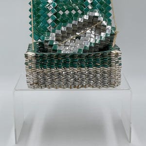 May include: A woven, rectangular box made of green and silver squares, with a matching lid. The box is made of recycled materials and is displayed on a clear acrylic stand. The lid is attached with twine.