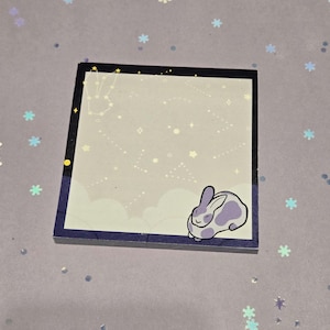 May include: Square notepad with a celestial design. The border is dark blue with a constellation and star pattern. The notepad features a light purple and white bunny illustration in the corner. The background is a light purple color.