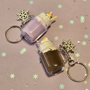 May include: Two mason jar keychain charms with a snowflake charm. One charm is filled with a purple milkshake with sprinkles and a straw. The other charm is filled with a chocolate milkshake with whipped cream and a straw.