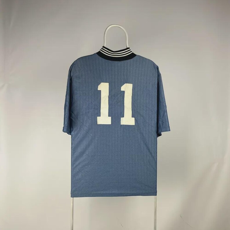 K&ouml;nnte beinhalten: Ein blaues Kurzarmtrikot mit der wei&szlig;en Nummer 11 auf dem R&uuml;cken. Der Kragen ist schwarz mit wei&szlig;en Streifen. Das Trikot h&auml;ngt an einem wei&szlig;en Kleiderb&uuml;gel.