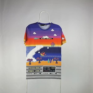 May include: A colorful t-shirt with a beach-themed graphic. The design features a sunset sky, palm trees, and surfers. Text includes "BEACH TROOPS", "TRICKS 0885", "JUMPS 1998", and other phrases. The shirt has a crew neck and short sleeves.
