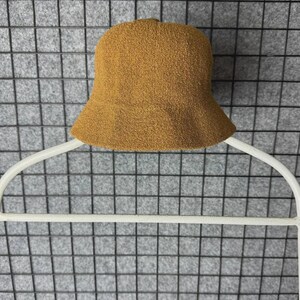 Puede incluir: Un sombrero de pescador de color marr&oacute;n con una superficie texturizada. El sombrero se muestra en una percha blanca sobre un fondo de rejilla gris. El ala del sombrero est&aacute; ligeramente ensanchada.