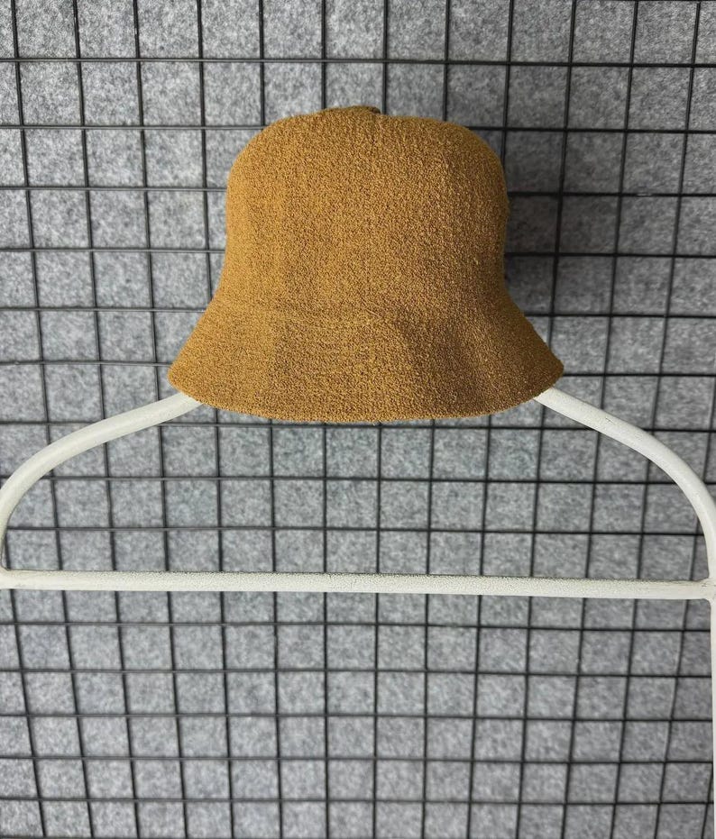 Puede incluir: Un sombrero de pescador de color marr&oacute;n con una superficie texturizada. El sombrero se muestra en una percha blanca sobre un fondo de rejilla gris. El ala del sombrero est&aacute; ligeramente ensanchada.