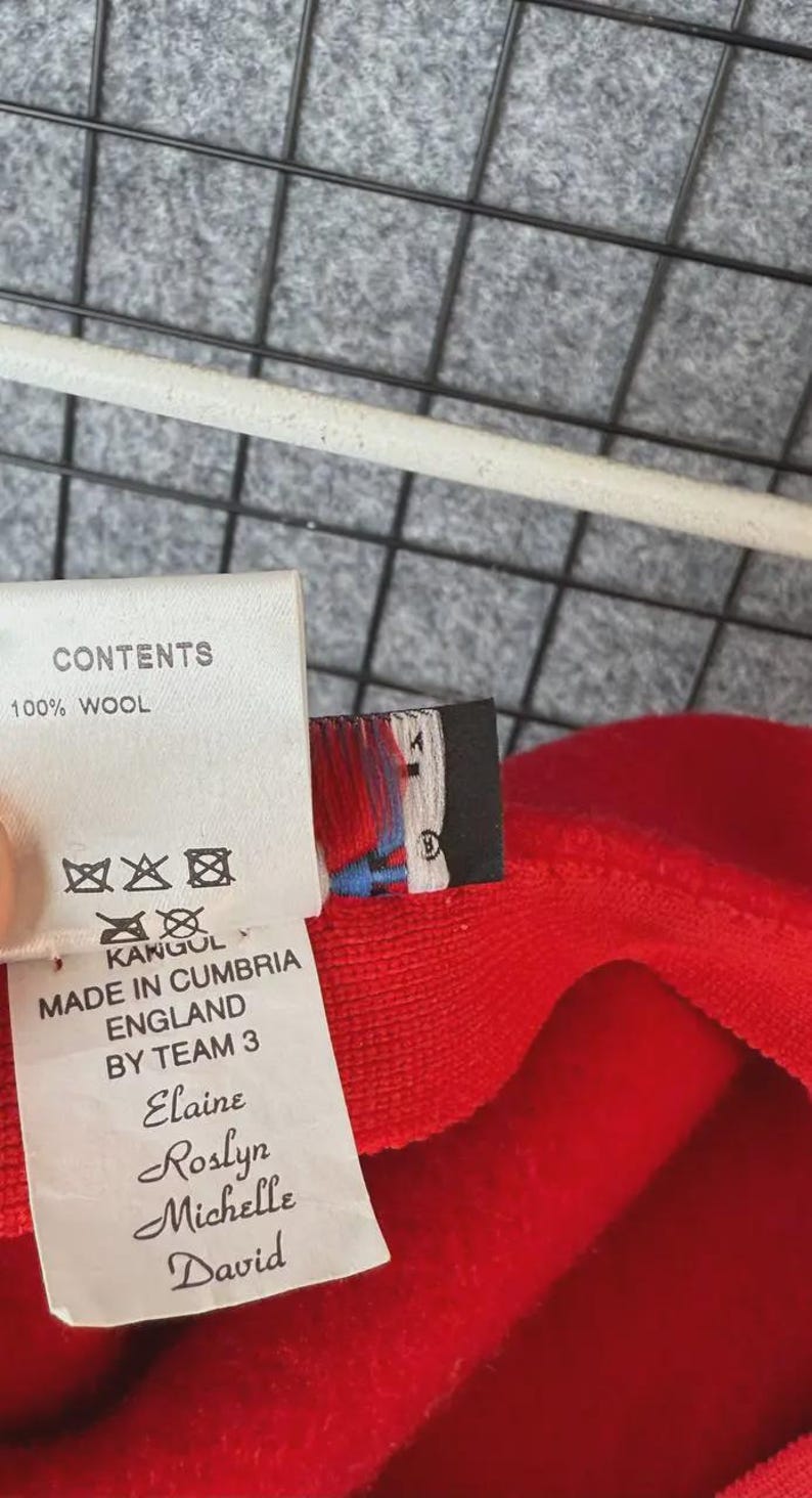 Puede incluir: Primer plano de una tela roja con una etiqueta. La etiqueta dice "CONTENTS 100% WOOL" y "MADE IN CUMBRIA ENGLAND BY TEAM 3". La marca "KANGOL" tambi&eacute;n es visible. La tela es de un rojo s&oacute;lido y vibrante.