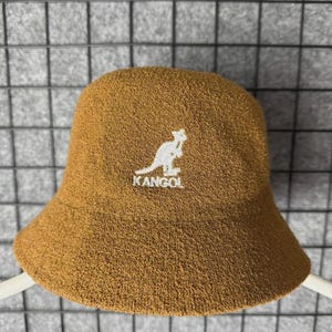 Puede incluir: Sombrero de cubo marr&oacute;n con el logotipo blanco de KANGOL con un canguro. El sombrero tiene un acabado suave y texturizado y un dise&ntilde;o cl&aacute;sico y redondeado. Un accesorio de moda para varios atuendos informales.