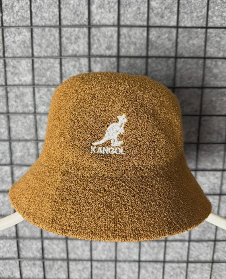 Puede incluir: Sombrero de cubo marr&oacute;n con el logotipo blanco de KANGOL con un canguro. El sombrero tiene un acabado suave y texturizado y un dise&ntilde;o cl&aacute;sico y redondeado. Un accesorio de moda para varios atuendos informales.