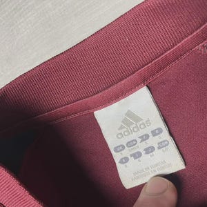 May include: Close-up of a maroon Adidas athletic shirt. The shirt's collar and tag are visible. The tag displays the Adidas logo and size information. The shirt is made in Tunisia.
