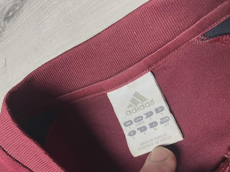 May include: Close-up of a maroon Adidas athletic shirt. The shirt's collar and tag are visible. The tag displays the Adidas logo and size information. The shirt is made in Tunisia.