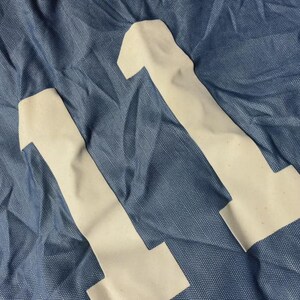 K&ouml;nnte beinhalten: Nahaufnahme eines blauen Stoffes mit zwei wei&szlig;en Applikationen der Zahl 11. Der Stoff hat ein strukturiertes Aussehen, und die Zahlen sind leicht cremefarben. Das Bild deutet auf ein Sporttrikot oder Sportbekleidung hin.