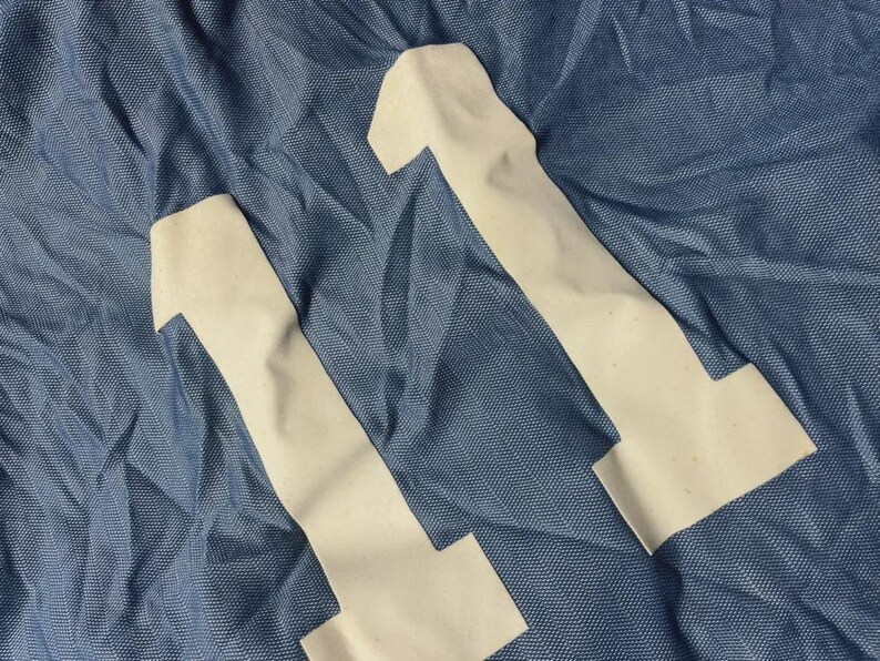 K&ouml;nnte beinhalten: Nahaufnahme eines blauen Stoffes mit zwei wei&szlig;en Applikationen der Zahl 11. Der Stoff hat ein strukturiertes Aussehen, und die Zahlen sind leicht cremefarben. Das Bild deutet auf ein Sporttrikot oder Sportbekleidung hin.