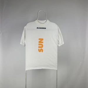 May include: White t-shirt with the text "JIL SANDER" in black at the top and the word "SUN" in orange vertically down the front. The shirt is hanging on a metal hanger against a gray background.