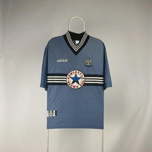 K&ouml;nnte beinhalten: Hellblaues Adidas-Fu&szlig;balltrikot mit einem schwarz-wei&szlig; gestreiften Design &uuml;ber der Brust. Das Trikot hat einen V-Ausschnitt mit schwarz-wei&szlig;en Zierleisten, das Adidas-Logo und ein Newcastle Brown Ale-Logo.