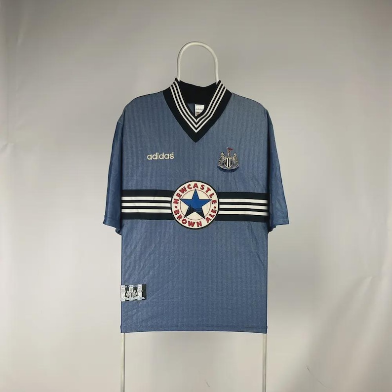 K&ouml;nnte beinhalten: Hellblaues Adidas-Fu&szlig;balltrikot mit einem schwarz-wei&szlig; gestreiften Design &uuml;ber der Brust. Das Trikot hat einen V-Ausschnitt mit schwarz-wei&szlig;en Zierleisten, das Adidas-Logo und ein Newcastle Brown Ale-Logo.