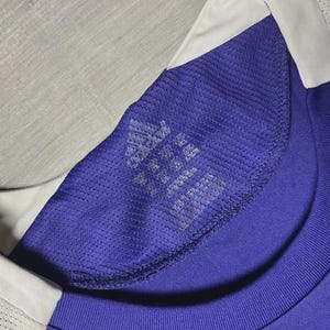 May include: Close-up of a blue athletic shirt with a white collar. The shirt has a mesh texture on the shoulder and a tag with the Adidas logo and text. The shirt is made of a breathable material.