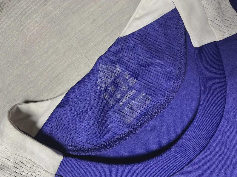 May include: Close-up of a blue athletic shirt with a white collar. The shirt has a mesh texture on the shoulder and a tag with the Adidas logo and text. The shirt is made of a breathable material.