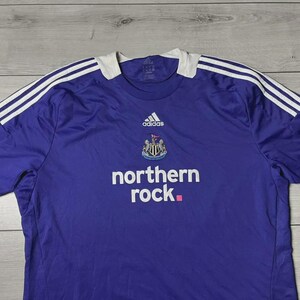 May include: A purple Newcastle United football shirt by Adidas. It has white stripes on the shoulders, a white collar, and the 'northern rock' logo. The Newcastle United crest and Adidas logo are also visible.