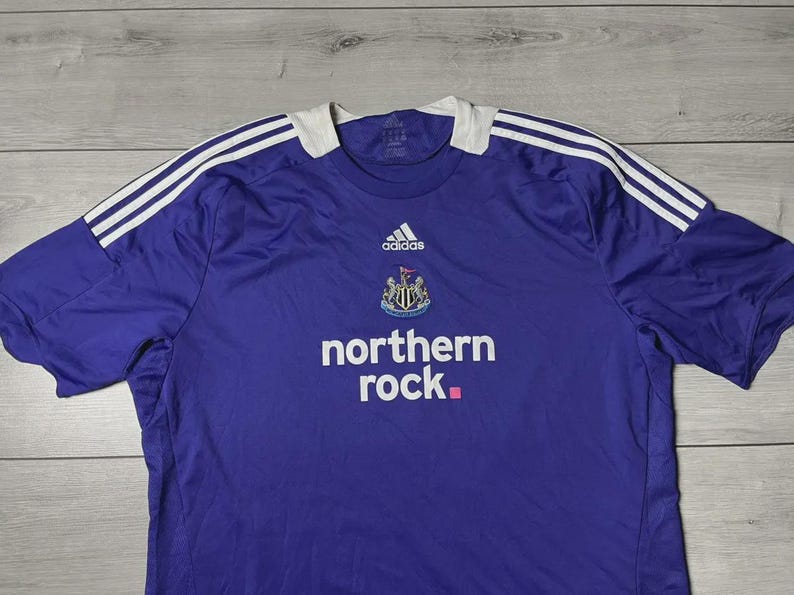 May include: A purple Newcastle United football shirt by Adidas. It has white stripes on the shoulders, a white collar, and the 'northern rock' logo. The Newcastle United crest and Adidas logo are also visible.