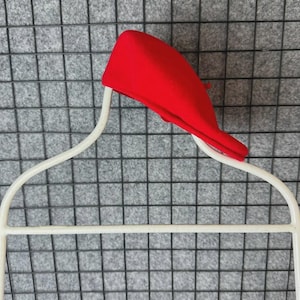 Puede incluir: Un boina roja brillante descansa sobre una percha de alambre blanco. La boina est&aacute; hecha de un material suave y est&aacute; en &aacute;ngulo, mostrando su forma. La percha tiene un dise&ntilde;o curvo, adecuado para guardar ropa.