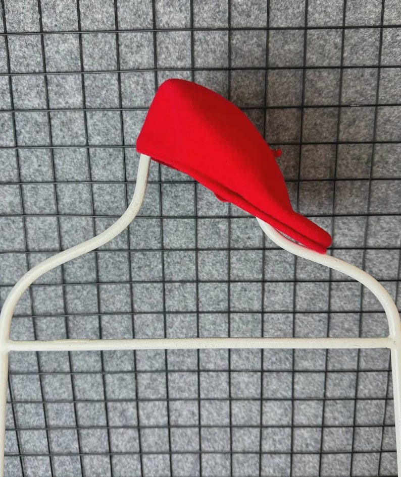 Puede incluir: Un boina roja brillante descansa sobre una percha de alambre blanco. La boina est&aacute; hecha de un material suave y est&aacute; en &aacute;ngulo, mostrando su forma. La percha tiene un dise&ntilde;o curvo, adecuado para guardar ropa.