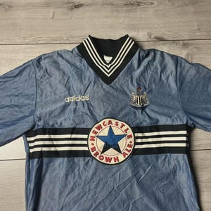 K&ouml;nnte beinhalten: Hellblaues Adidas-Fu&szlig;balltrikot mit schwarz-wei&szlig; gestreiftem Kragen und schwarzen Streifen auf der Brust. Das Trikot zeigt das Wappen von Newcastle United und einen Aufn&auml;her "Newcastle Brown Ale".