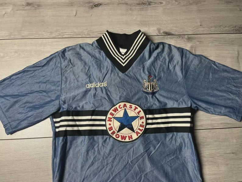 K&ouml;nnte beinhalten: Hellblaues Adidas-Fu&szlig;balltrikot mit schwarz-wei&szlig; gestreiftem Kragen und schwarzen Streifen auf der Brust. Das Trikot zeigt das Wappen von Newcastle United und einen Aufn&auml;her "Newcastle Brown Ale".