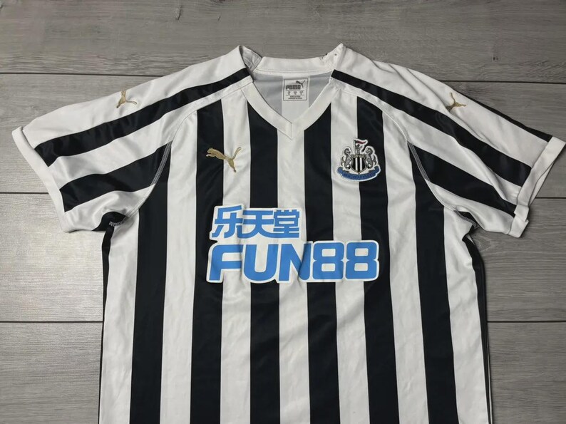 Puede incluir: Camiseta de f&uacute;tbol a rayas blancas y negras con cuello en V. Presenta el escudo del Newcastle United, el logotipo de Puma y el logotipo del patrocinador Fun88 en azul. La camiseta est&aacute; hecha de un material ligero.