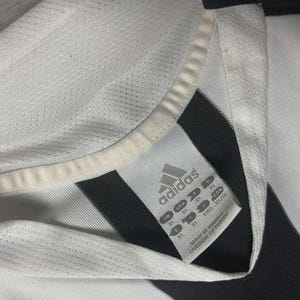Pu&ograve; includere: Primo piano di una maglia Adidas bianca e nera. La maglia ha un colletto bianco e un'etichetta con il logo Adidas e informazioni sulla taglia. La maglia &egrave; realizzata in un tessuto traspirante e strutturato.