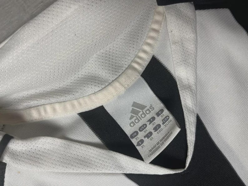 Pu&ograve; includere: Primo piano di una maglia Adidas bianca e nera. La maglia ha un colletto bianco e un'etichetta con il logo Adidas e informazioni sulla taglia. La maglia &egrave; realizzata in un tessuto traspirante e strutturato.
