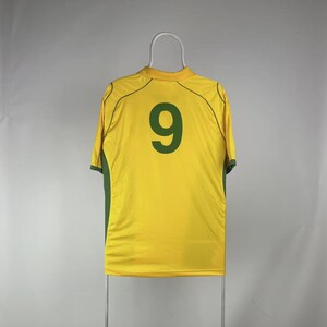 May include: A yellow athletic jersey with green accents and the number 9 in dark green on the back. The jersey is hanging on a white hanger against a plain background. The jersey has short sleeves and a crew neck.