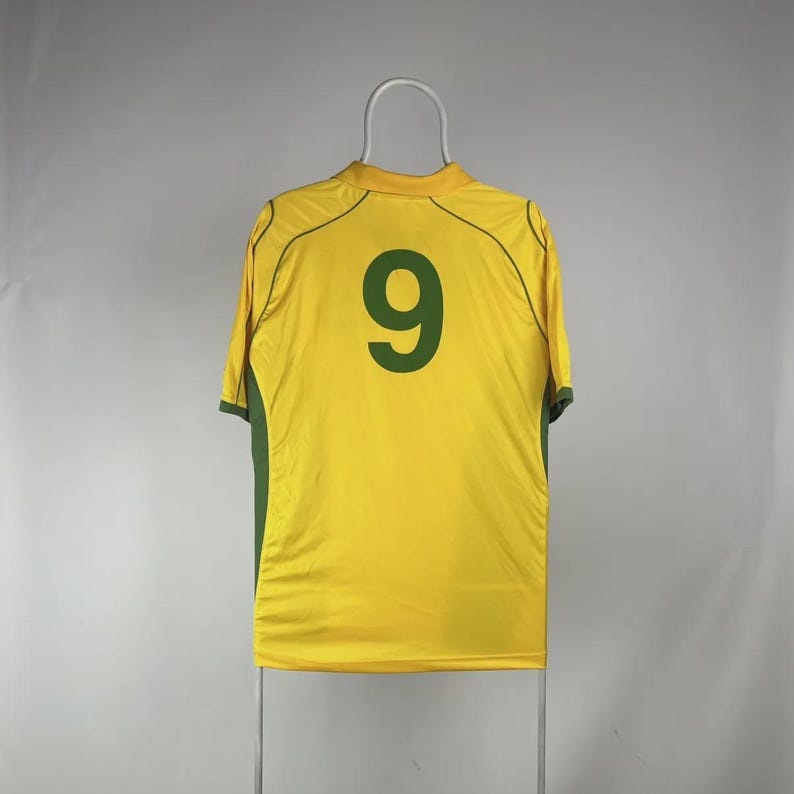 May include: A yellow athletic jersey with green accents and the number 9 in dark green on the back. The jersey is hanging on a white hanger against a plain background. The jersey has short sleeves and a crew neck.