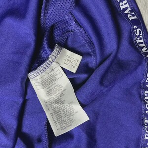 May include: Close-up of a purple athletic jersey with white text along the side that reads "EST 1892 ST JAMES' PARK". The jersey has a tag with the Adidas logo and product information. The jersey is made of a polyester blend.