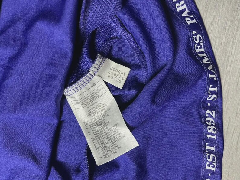 May include: Close-up of a purple athletic jersey with white text along the side that reads "EST 1892 ST JAMES' PARK". The jersey has a tag with the Adidas logo and product information. The jersey is made of a polyester blend.