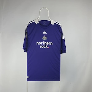 May include: A blue football shirt with white detailing and the words "northern rock." The Adidas logo and a team crest are visible. The shirt is displayed on a white hanger.