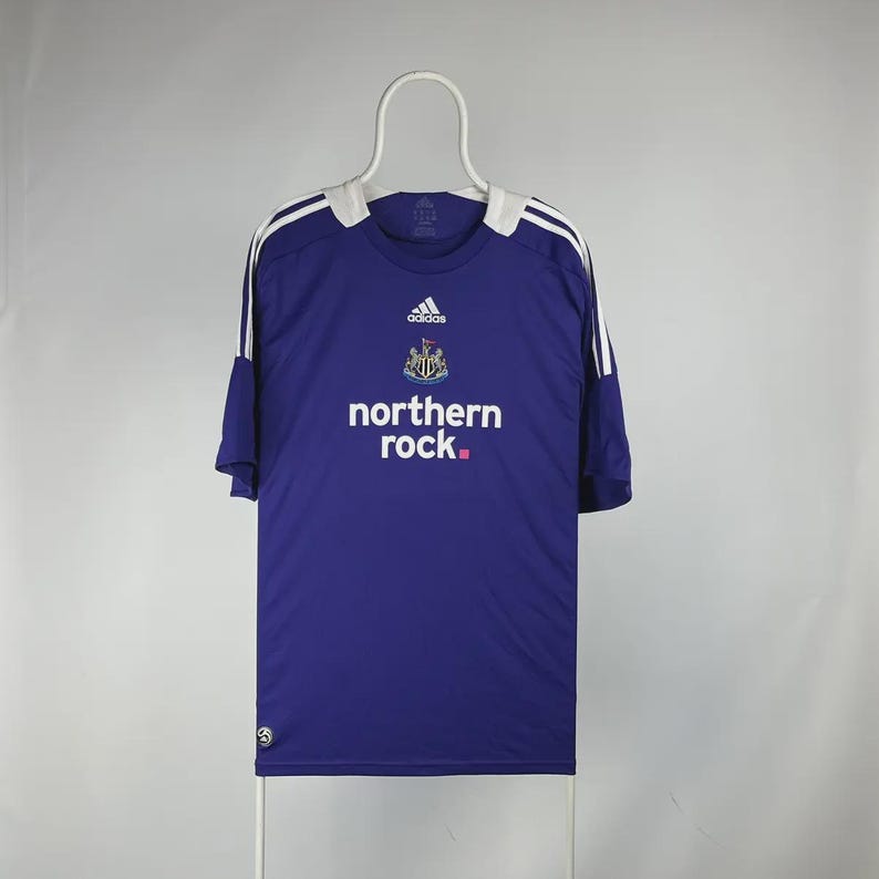 May include: A blue football shirt with white detailing and the words "northern rock." The Adidas logo and a team crest are visible. The shirt is displayed on a white hanger.