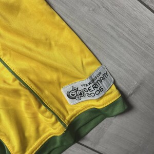 May include: Close-up of a yellow and green soccer jersey. The jersey has a green trim and a patch that reads "FIFA WORLD CUP GERMANY 2006". The jersey is made of a shiny, athletic material.
