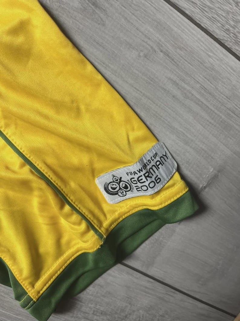 May include: Close-up of a yellow and green soccer jersey. The jersey has a green trim and a patch that reads "FIFA WORLD CUP GERMANY 2006". The jersey is made of a shiny, athletic material.